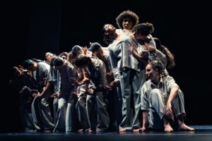 eine Gruppe Tänzerinnen und Tänzer in heller Kleidung, mit den Körpern eine aufsteigende Linie bildend