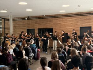 Ein Kammerorchester spielt im Stehen vor einer Gruppe Schüler.