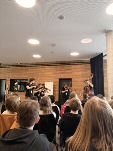 Fünf Musikerinnen und Musiker mit Streichinstrumenten spielen in einer Schulaula vor Schülern.