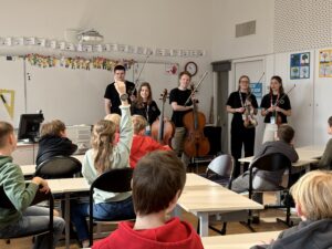 Fünf junge Musikerinnen udn Musiker mit Streichinstrumenten stehen vor einer Klasse im Klassenzimmer.