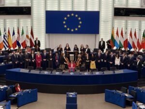 Plenarsaal des Europäischen Parlaments; eine Gruppe Musiker im Hintergrund; darüber die Europafahne. Eine Frau im roten Jackett vorne am Rednerpult