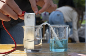 Experiment. Zwei Erlenmeierkolben; im linken klare Flüssigkeit, Metallplatte mit Kabel; im rechten blaue Flüssikeit.