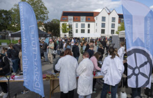 Ein großer Platz mit Ständen; Besucher, im Vordergrund junge Menschen in Laborkitteln.
